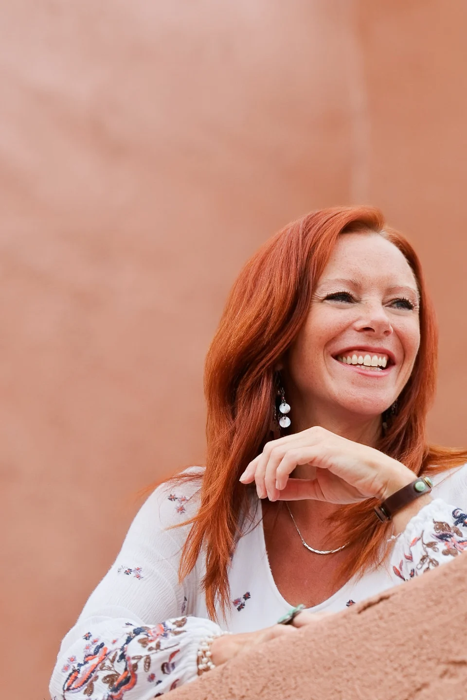 Angela smiling in a white embroidered dress against a warm stucco wall